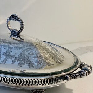 Op de afbeelding: Een ovale serveerschaal van zilver met een glazen deksel en sierlijke handvatten. De schaal heeft een decoratief gegraveerd ontwerp en staat op drie poten.