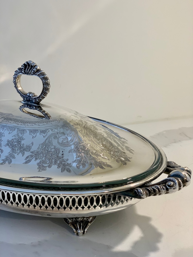 Op de afbeelding: Een ovale serveerschaal van zilver met een glazen deksel en sierlijke handvatten. De schaal heeft een decoratief gegraveerd ontwerp en staat op drie poten.