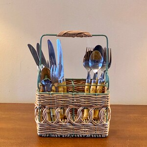 May include: A wicker cutlery caddy with a green metal frame and handle, holding various stainless steel knives, forks, and spoons. The caddy features bamboo-style accents and a woven design, perfect for kitchen or dining use.