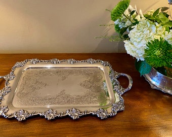 May include: A silver serving tray with ornate grapevine detailing around the edge and two handles. The tray has a slightly raised rim and a smooth, reflective surface.