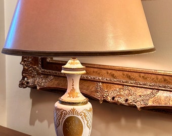 May include: A white ceramic table lamp with gold accents and a floral design. The lamp has a beige fabric shade.