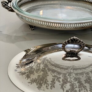 Op de afbeelding: Zilveren serveerschaal met een glazen deksel. De schaal heeft een decoratief bloemmotief en een gegolfde rand. Het deksel heeft een decoratieve knop.