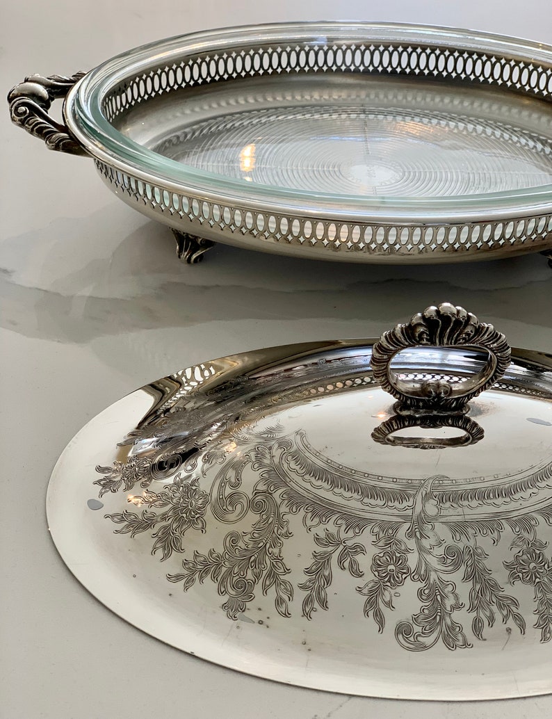Op de afbeelding: Zilveren serveerschaal met een glazen deksel. De schaal heeft een decoratief bloemmotief en een gegolfde rand. Het deksel heeft een decoratieve knop.