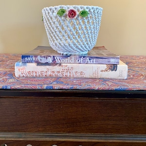 May include: Two books stacked on a wooden table with a white ceramic basket with a pink rose and green leaves on top. The top book is titled "The Renaissance" and the bottom book is titled "A World of Art".