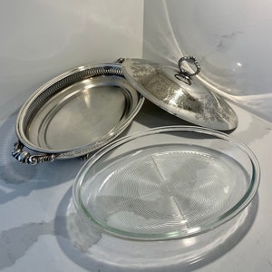 Op de afbeelding: Een ovale serveerschaal van zilver met een glazen inzet en een bijpassend deksel. De schaal heeft een decoratieve rand en sierlijke handvatten.