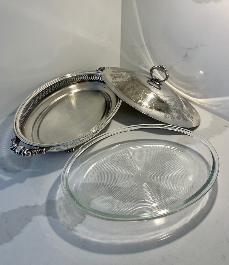 Op de afbeelding: Een ovale serveerschaal van zilver met een glazen inzet en een bijpassend deksel. De schaal heeft een decoratieve rand en sierlijke handvatten.