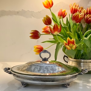 Op de afbeelding: Een zilveren ovale serveerschaal met een deksel en een bloemenpatroon. De schaal staat op een wit oppervlak en er zijn oranje en gele tulpen op de achtergrond.