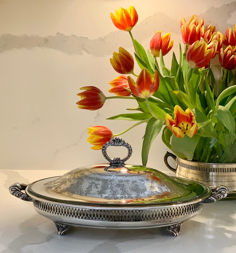 Op de afbeelding: Een zilveren ovale serveerschaal met een deksel en een bloemenpatroon. De schaal staat op een wit oppervlak en er zijn oranje en gele tulpen op de achtergrond.