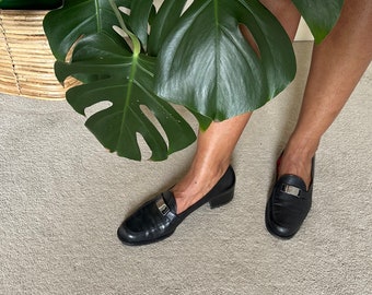 Puede incluir: Un par de mocasines de cuero negro con hebillas plateadas. Los mocasines tienen un tacón bajo y los lleva una persona con las piernas descubiertas.