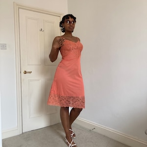 May include: A coral pink slip dress with lace trim. The dress has a fitted bodice and a flowing skirt. The model is wearing the dress with white sandals.
