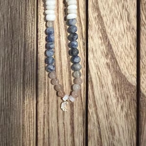 Peut inclure: Un collier de perles avec des perles blanches, bleues et roses. Le collier comporte un charm en forme de croissant de lune. Les perles sont enfilées sur un cordon, sur un fond en bois.