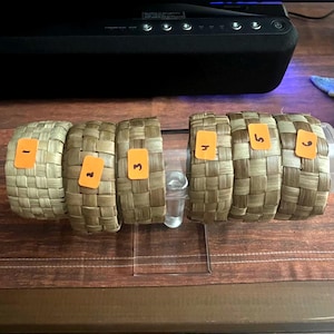 May include: Six woven bands arranged in two groups of three, each featuring a light to medium brown natural fiber texture. Each band has an orange rectangular sticker attached, numbered one through six in black ink, displayed on a clear stand.