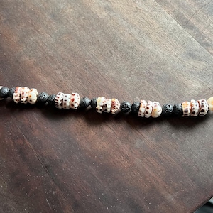 May include: A necklace featuring alternating sections of small, patterned seashells and black, round lava beads. The seashells have a cream base with brown and red markings. The necklace is displayed on a dark wooden surface.