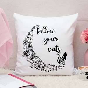 以下が含まれることがあります： 黒い文字で「Follow your cats」と書かれた白いクッション。花の模様がついた黒い猫のシルエットが描かれている。猫は花のつるでできた三日月型の中に座っている。