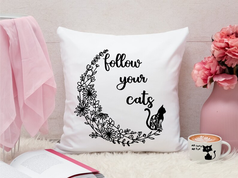以下が含まれることがあります： 黒い文字で「Follow your cats」と書かれた白いクッション。花の模様がついた黒い猫のシルエットが描かれている。猫は花のつるでできた三日月型の中に座っている。