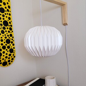 Pu&ograve; includere: Un lampadario bianco in stile origami &egrave; appeso a un supporto a parete in legno. La luce &egrave; sospesa da un cavo bianco e ha una forma sferica plissettata.