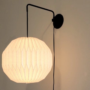 May include: A white, pleated, round lampshade hangs from a black metal arm attached to a wall-mounted base. The lampshade is lit and casts a warm glow.