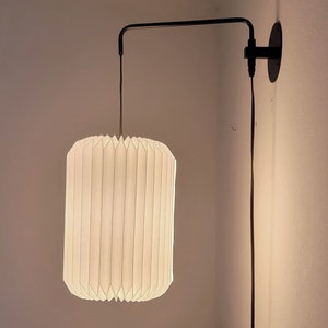 May include: A white paper lampshade with a pleated design hangs from a black metal arm attached to a wall. The lampshade is illuminated, casting a warm glow.