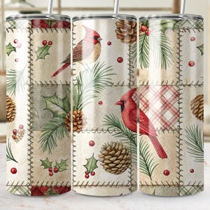 May include: A stainless steel tumbler with a patchwork design featuring red cardinals, pinecones, holly, and plaid patterns. The tumbler has a straw and is decorated with a winter theme.