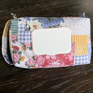May include: A patchwork wipe case featuring a variety of floral and gingham patterns in shades of pink, blue, yellow, and green. The case has a white plastic dispenser lid and a small fabric handle.