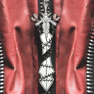 Könnte beinhalten: Ein silberner und weißer Emaille-Pin in Form eines Schwertes mit einem Schädel- und Rippenkorb-Design. Der Pin ist auf einer roten Lederjacke mit einem schwarzen Reißverschluss angebracht.