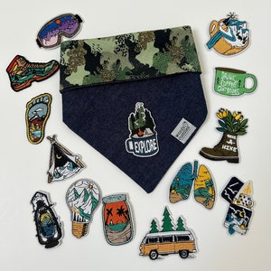 May include: A collection of embroidered iron-on patches featuring outdoor and nature themes. The patches include a camping tent, a lightbulb with mountains, a campfire mug, a hiking boot, a coffee cup, and a camper van. The patches are on a white background.