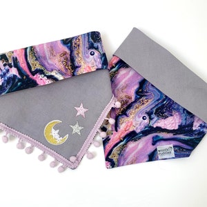 May include: Purple, pink and gold marbled dog bandana with a grey felt backing. The bandana has a pink pom-pom trim and features a gold crescent moon and silver stars. The bandana has a label that says "woof cuddles".