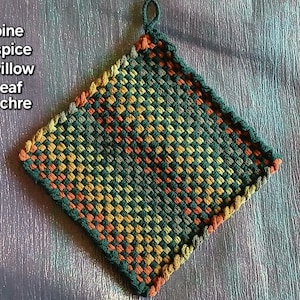 May include: A square woven potholder featuring a checkered pattern in shades of pine, spice, willow, leaf, and ochre. The text 'pine spice willow leaf ochre' is printed on the left side. It includes a small loop at the top corner.