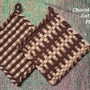 May include: Two square potholders featuring a woven checkerboard pattern in shades of dark brown, light brown, and tan. Each potholder has a small loop at the top corner for hanging. The text Chocolate Autumn Flax is printed in white on the right.