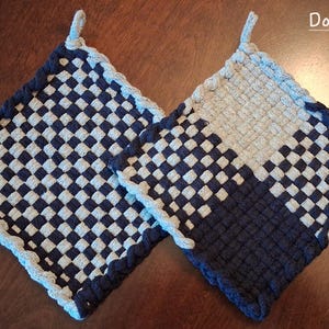 May include: Two square potholders made of woven fabric. One features a dark navy and silver checkered pattern. The other has a color-blocked design with sections of solid dark navy, solid silver, and a checkered pattern. Each has a small hanging loop.