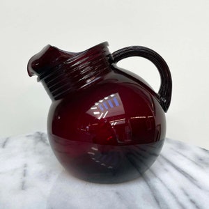 May include: A dark red glass pitcher with a round body and a handle. The pitcher is made of a thick, heavy glass and has a glossy finish.