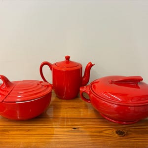 May include: Three-piece red ceramic cookware set. Includes a lidded bowl, a teapot, and a lidded casserole dish. The set is glossy and sits on a wooden surface. The bowl has a large handle, and the casserole dish has two side handles.