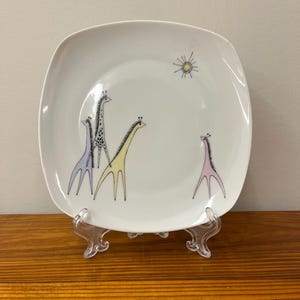 May include: A white square ceramic plate with a stylized sun design and four giraffes in different colors: blue, yellow, black and pink.