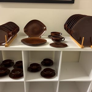 May include: A collection of brown ceramic dishware, including plates, saucers, and cups, displayed on a white shelf. The plates are square with rounded corners. The cups and saucers are small and round. The dishware is arranged in sets.