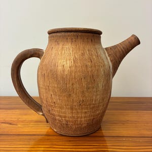 May include: A brown ceramic teapot with a lid, handle, and spout. The teapot has a textured surface with vertical lines and a light brown color. The handle is curved, and the spout extends from the side. The teapot is likely used for brewing tea.