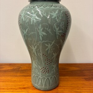 May include: A celadon-colored ceramic vase with a flared base and a narrow neck. The vase is decorated with a repeating pattern of vines, leaves, and clusters of red berries. The vase is displayed on a wooden surface.