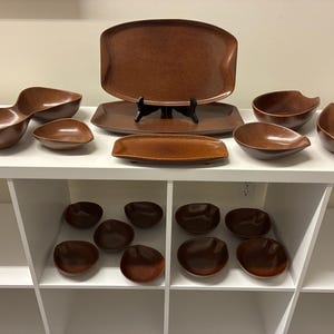 May include: A collection of brown ceramic serving dishes and bowls. The set includes a large oval platter, a rectangular tray, and various sized bowls. The pieces are arranged on a white shelf, showcasing their varied shapes and sizes.