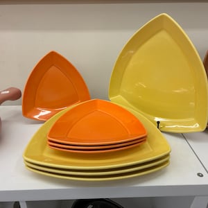 May include: A collection of colorful, triangular-shaped plates in shades of yellow and orange. The plates are stacked and arranged on a white surface, showcasing a modern and vibrant aesthetic. The plates are made of ceramic.