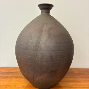 May include: A dark brown, matte-finished ceramic vase with a rounded, bulbous body and a narrow, flared neck. The surface features subtle horizontal ridges and a distinct vertical seam running down the center, with faint reddish-brown undertones.