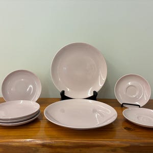 May include: A collection of light pink ceramic plates and saucers. The set includes various sizes of plates, including dinner plates, salad plates, and saucers. The plates are arranged on a wooden surface.