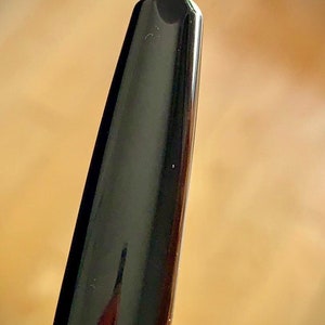 May include: Close-up of a black fountain pen nib with a gold trim. The nib is pointed and has a fine tip.