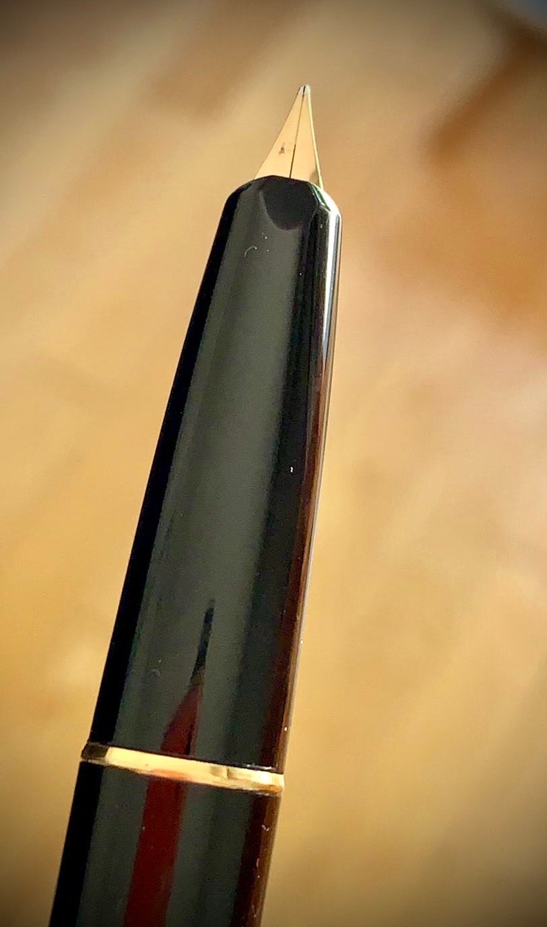 May include: Close-up of a black fountain pen nib with a gold trim. The nib is pointed and has a fine tip.