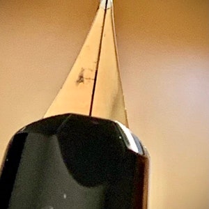 May include: Close-up of a gold-toned fountain pen nib. The nib is pointed and has a fine tip.