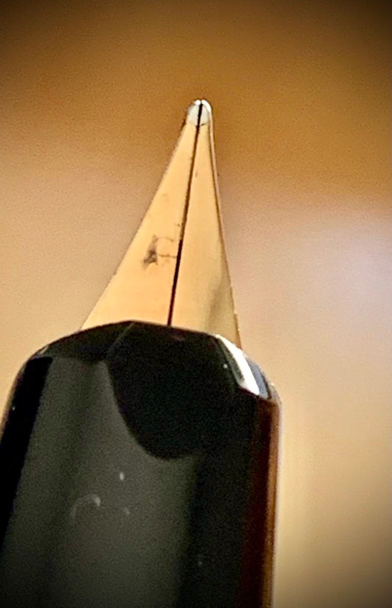 May include: Close-up of a gold-toned fountain pen nib. The nib is pointed and has a fine tip.