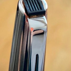 May include: Close-up of the top of a silver pen with a black rubber grip. The pen has a grooved design.