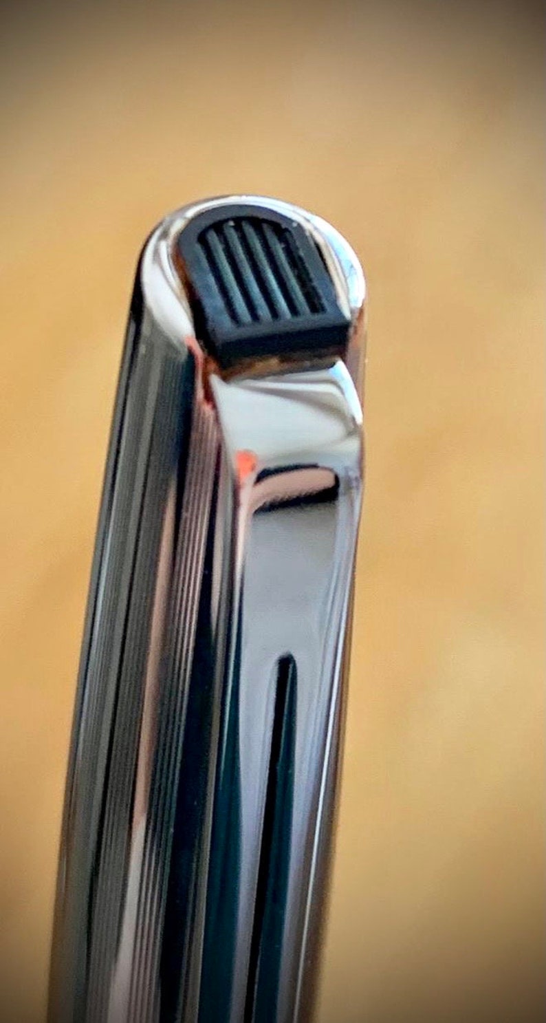 May include: Close-up of the top of a silver pen with a black rubber grip. The pen has a grooved design.