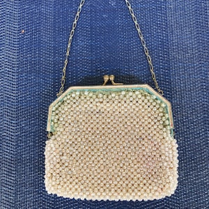 Puede incluir: Un bolso de noche de cuentas blancas con una correa de cadena dorada. El bolso está hecho de pequeñas cuentas redondas que están cosidas juntas en un patrón de cuadrícula. El bolso tiene un cierre de broche y una pequeña forma rectangular.