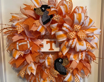 May include: Orange and white wreath with a Tennessee Volunteers theme. The wreath features a football, a large "T" and two black and gold dog silhouettes. The wreath is decorated with orange and white ribbons and mesh.