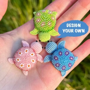 May include: Three crocheted turtle figurines in green, pink, and blue, each with a unique floral pattern. The green turtle has a beaded texture. A blue speech bubble with the text "DESIGN YOUR OWN" is also visible.