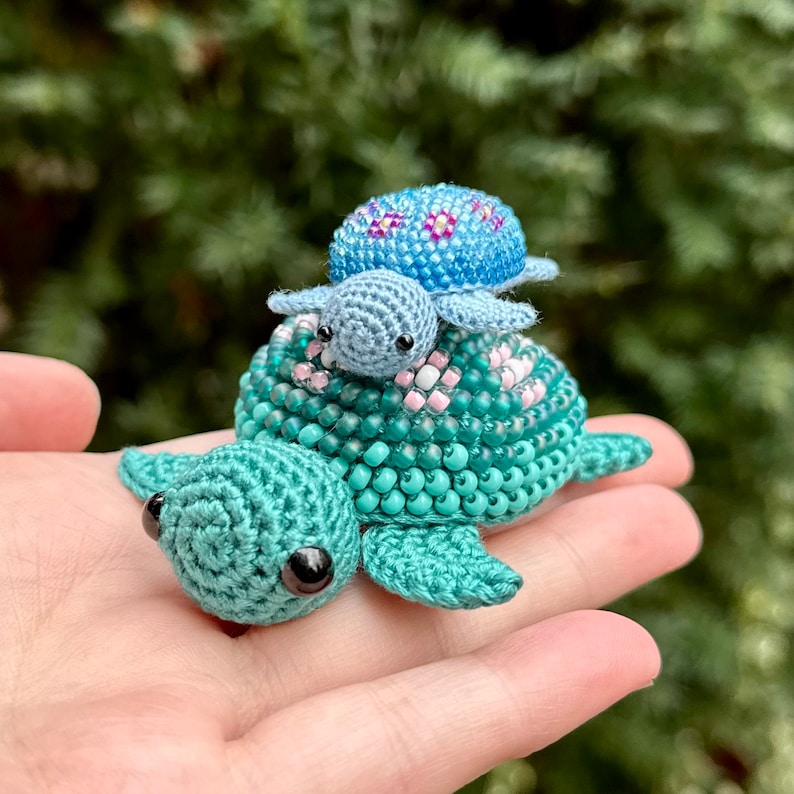 May include: A handmade crochet turtle family in shades of teal, blue, and pink. The largest turtle is teal with pink bead accents, supporting a smaller blue turtle with a beaded shell, and a tiny blue turtle on top.